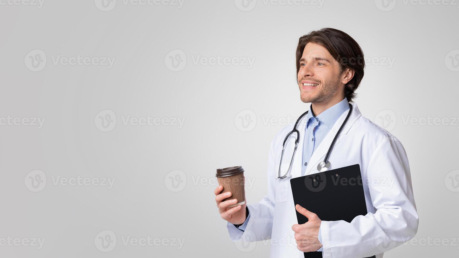 Happy young doctor holding clipboard with medical info and having coffee break over light studio background, free space photo