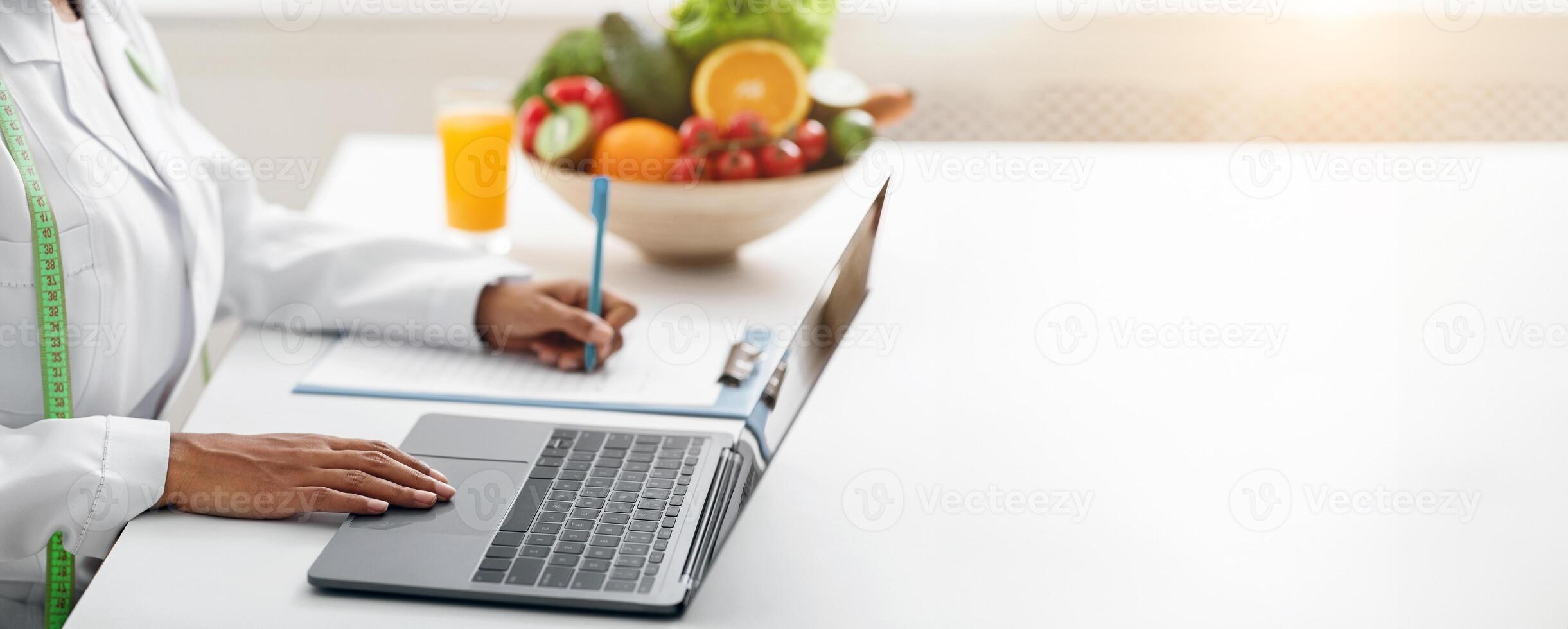 Cropped of black woman nutritionist working online, using laptop and writing down client history, clinic interior photo