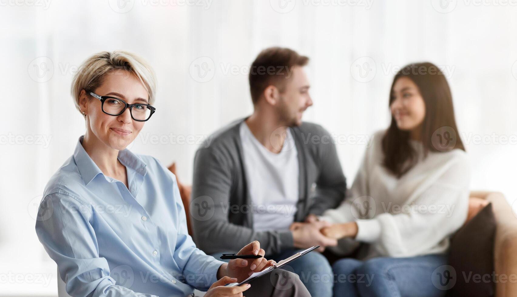 Professional Family Psychologist Smiling To Camera While Her Patients Couple Reconciling Sitting On Sofa In Office. Saved Marriage Concept photo