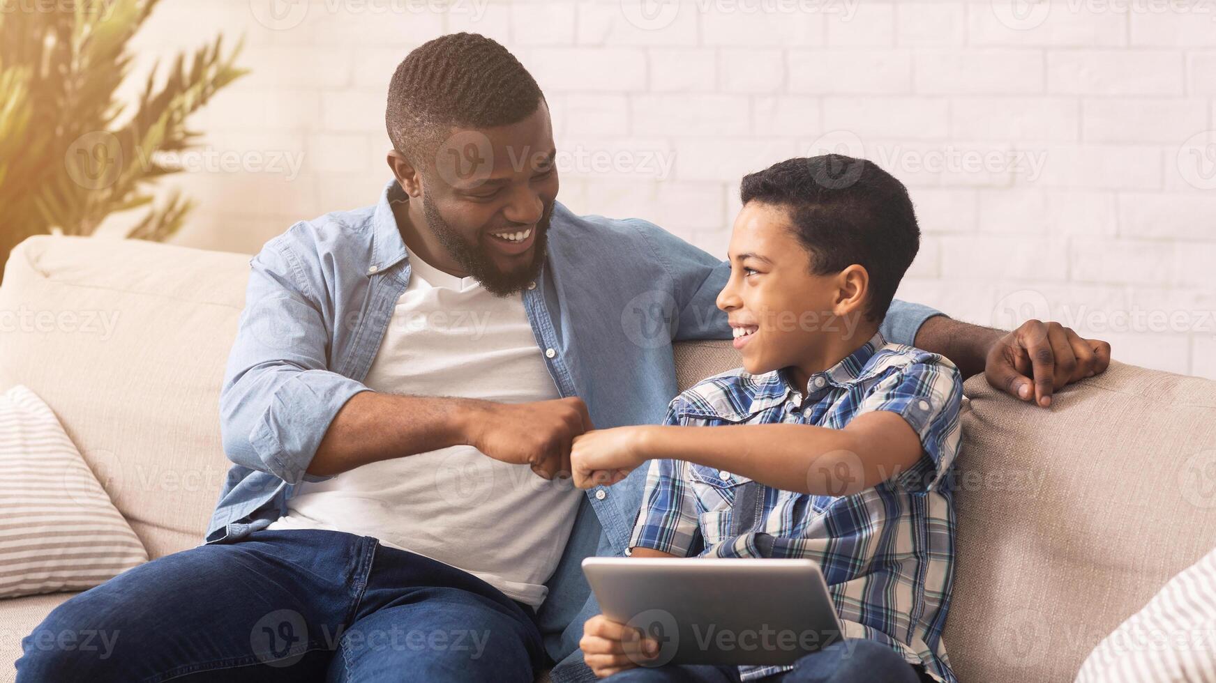 Joyful afro father and son bumping fists, celebrating online game victory with digital tablet, spending time together at home photo