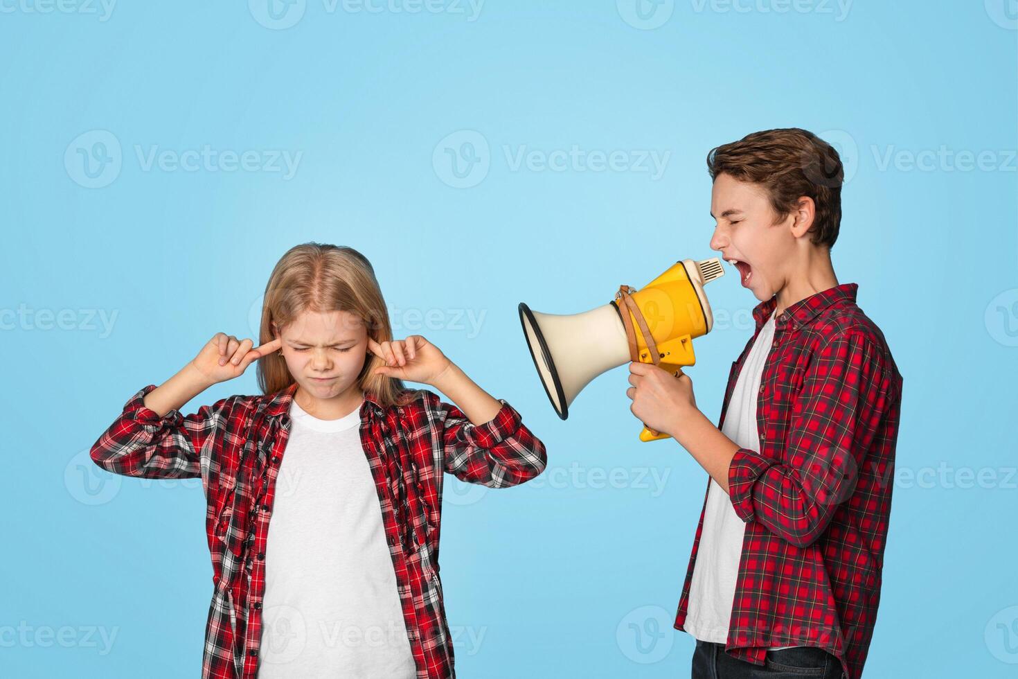 Misunderstanding Between Siblings. Little girl closing ears with fingers while her elder brother shouting to megaphone, blue background photo