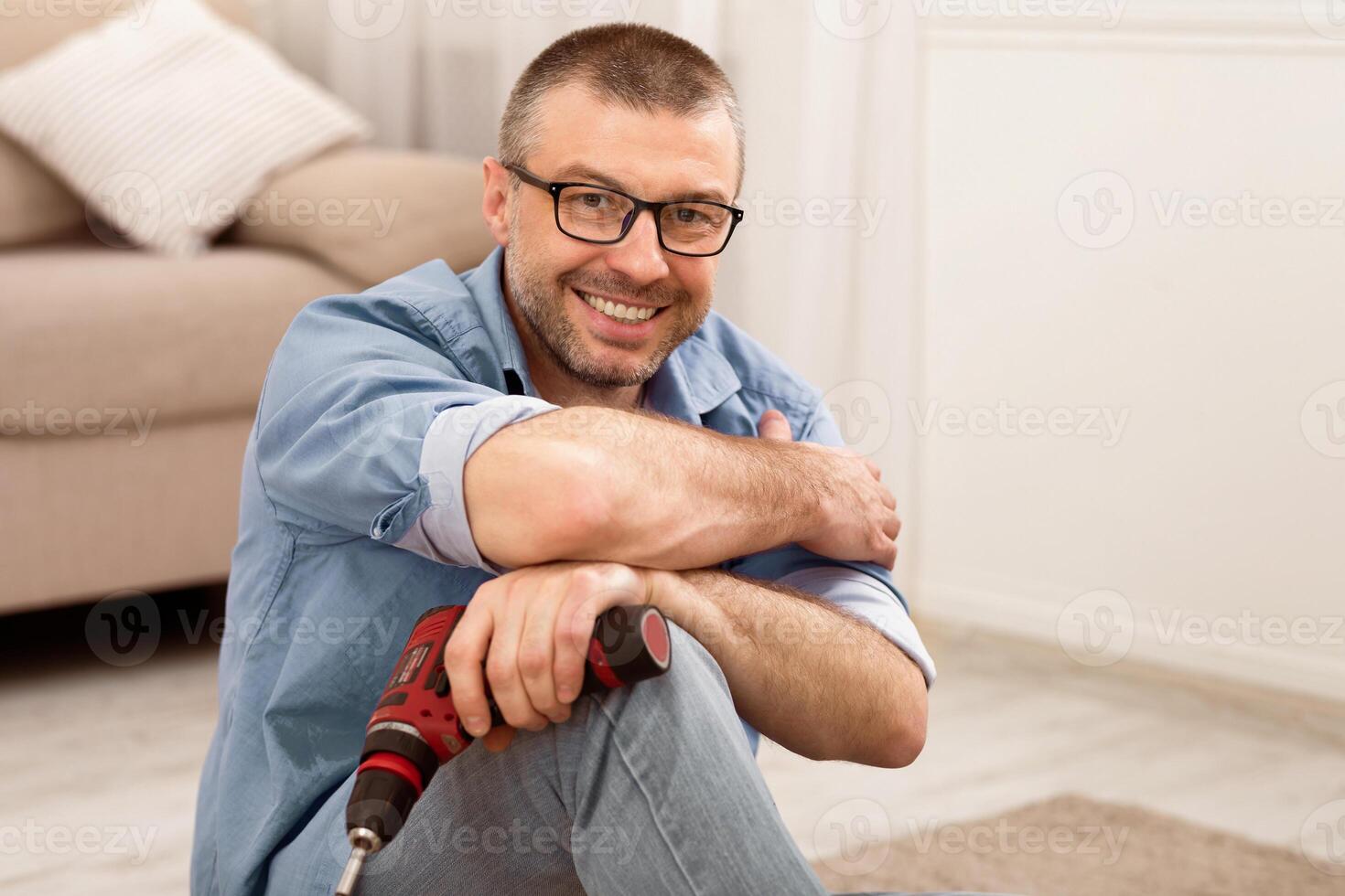 Mature Man Assembling Furniture Sitting Posing With Electric Drill, Smiling To Camera At Home. Carpentry Concept photo