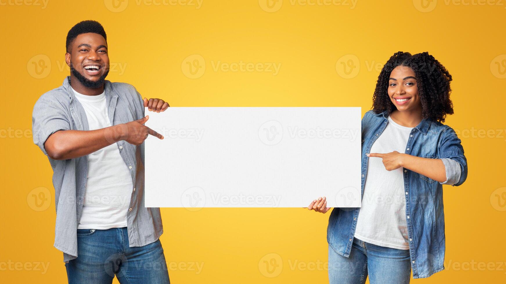Place For Your Ad. Cheerful black couple holding and pointing at blank advertisement placard, standing together on yellow background, copy space photo