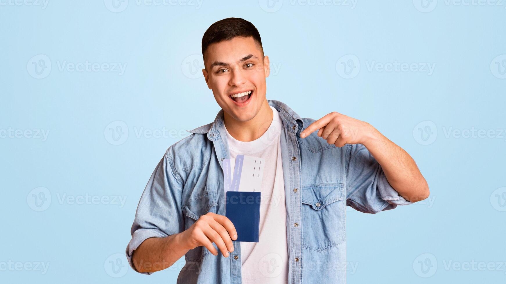 Cheery young guy pointing at passport and flight tickets in his hand over yellow photo