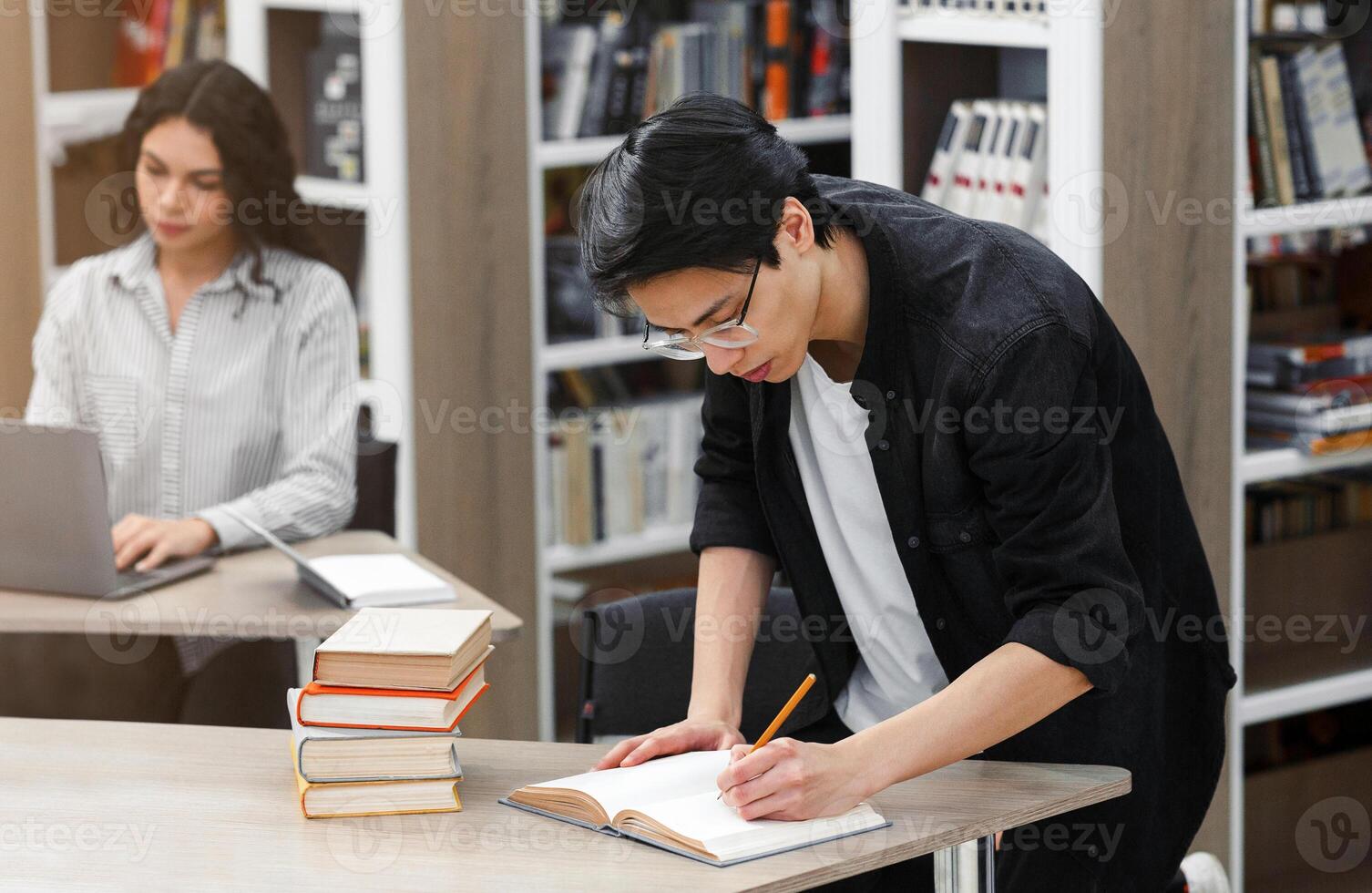Educational Process. Asian guy writing in notebook, standing at desk in campus library, latin student in the background photo