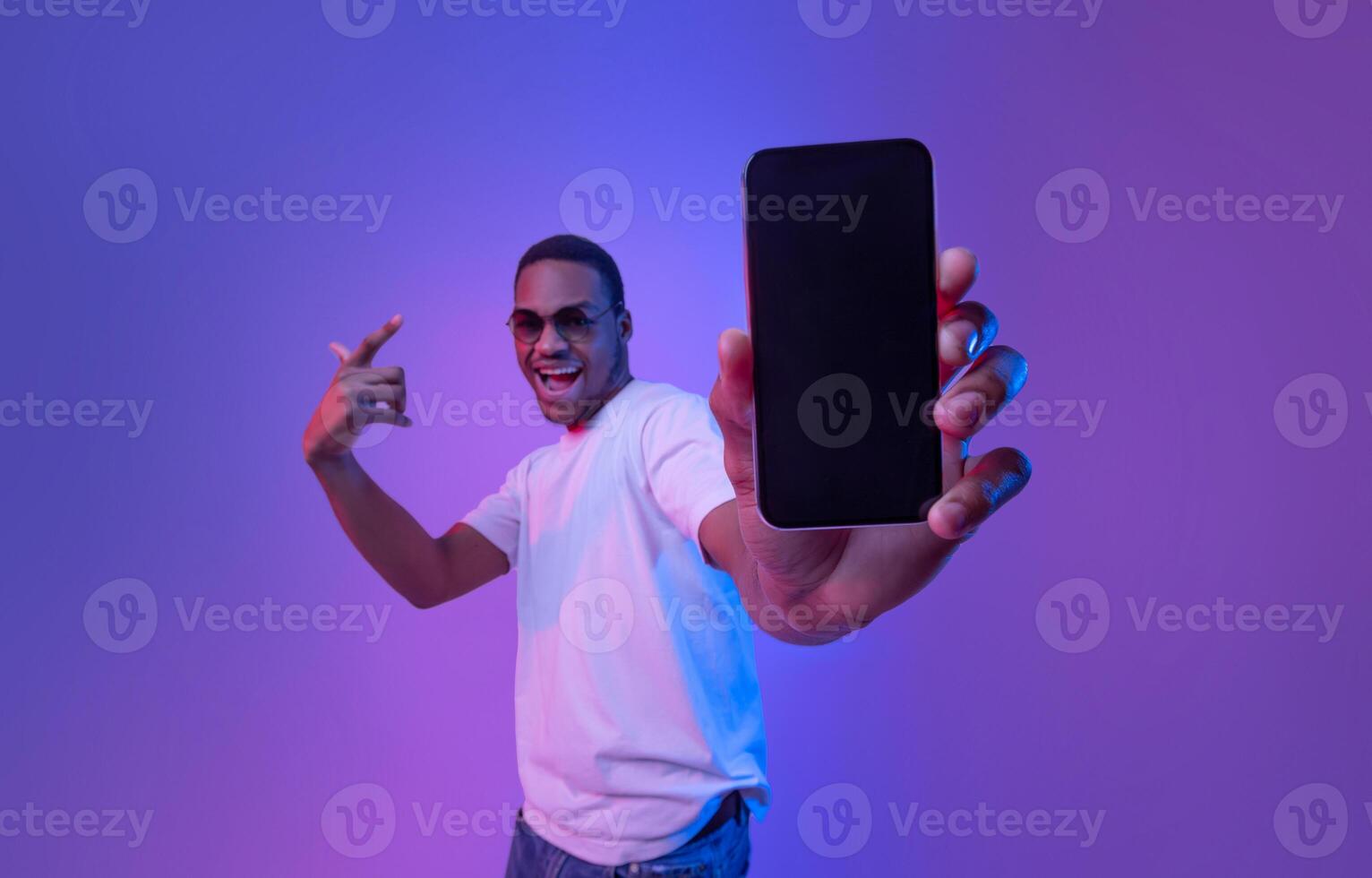 Cool Mobile Offer. Excited African Guy Demonstrating Smartphone With Black Screen, Standing Under Neon Lighting, Joyful Millennial Man Showing Free Mockup Copy Space For Your App Or Website Design photo
