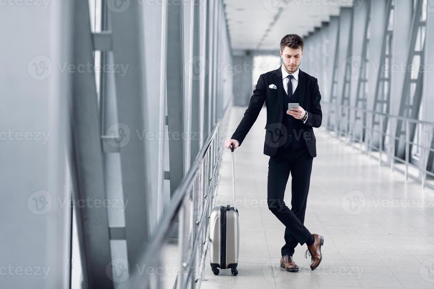 Portrait of confident guy in suit using cell phone standing in airport lobby holding trolley bag, copy space, full length photo