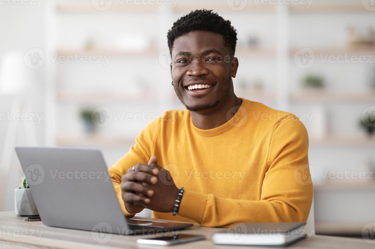 Portrait of positive young black guy in yellow sweater sitting at table, working on laptop, sending emails, office interior. Cheerful african american man freelancer working from home, copy space photo