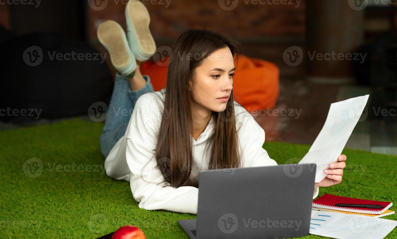 Freelancer is lying on floor, looking at document, works with laptop and notepad. Free space photo
