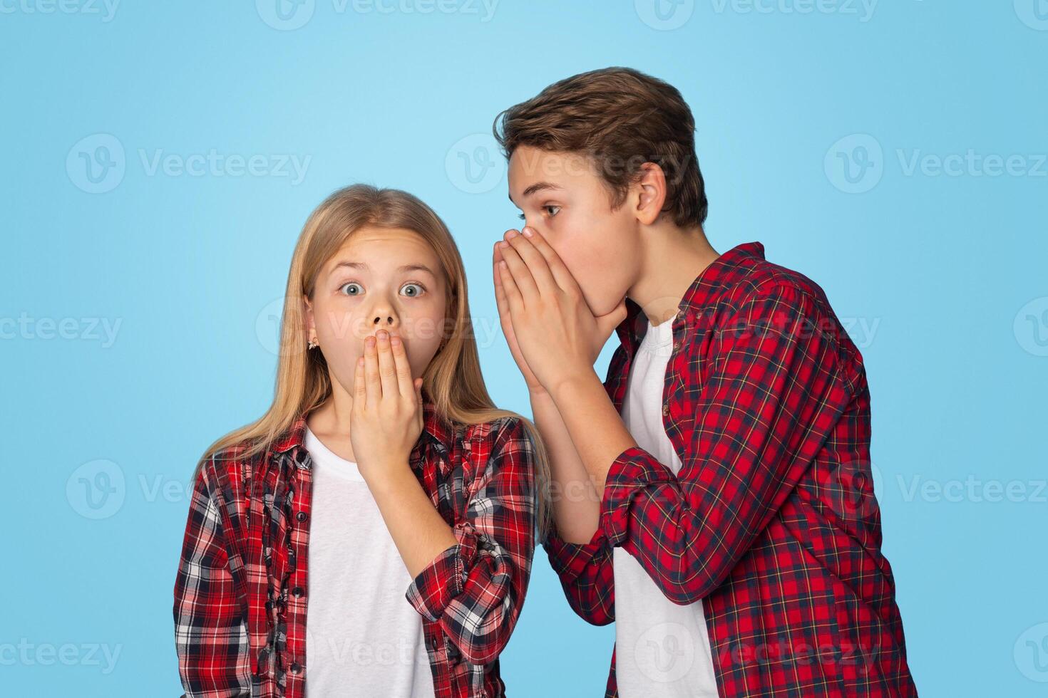 Teen Brother Sharing Secrets With His Little Sister, Whispering Gossips Into Her Ear, Standing Together On Blue Background With Copy Space photo