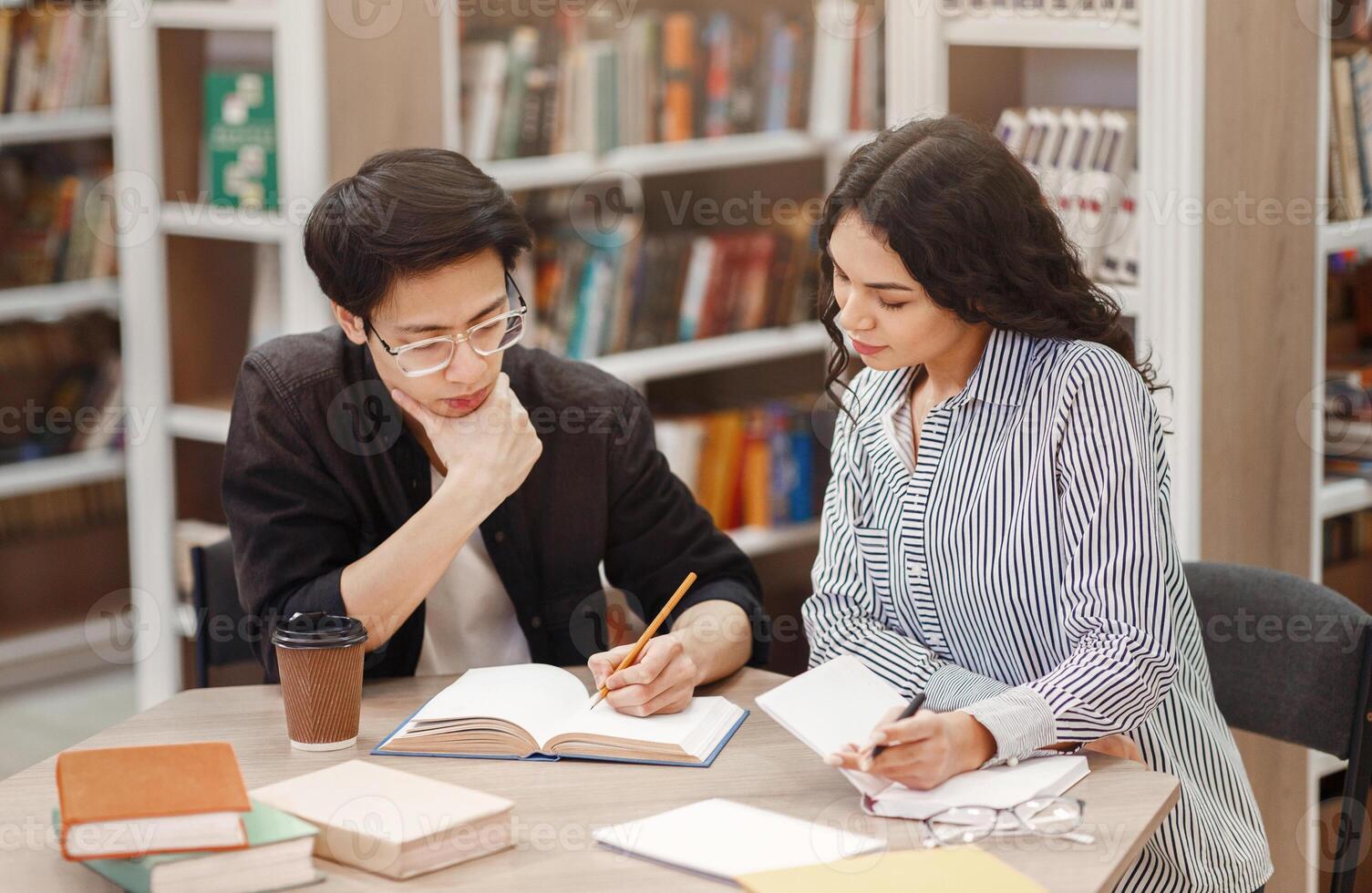 Studying Together. Concentrated multicultural couple doing homework, helping each other sitting at table in library photo