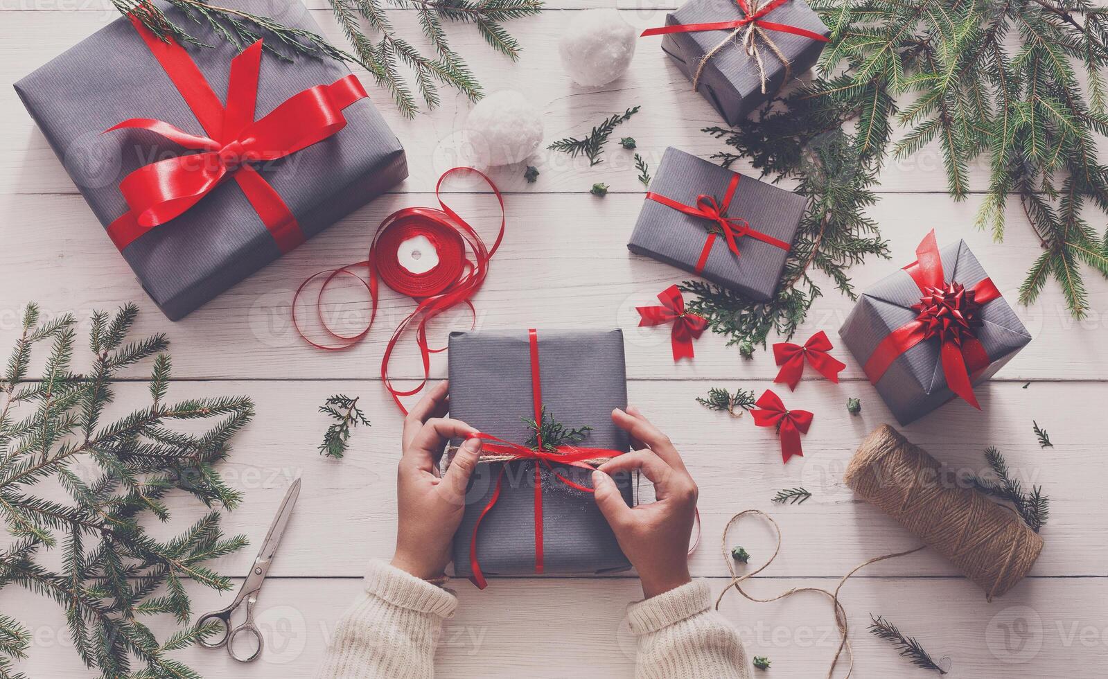Creative hobby. Gift wrapping. Packaging modern christmas present boxes in stylish gray paper with satin red ribbon. Top view of hands on white wood table with fir tree branches, decoration photo