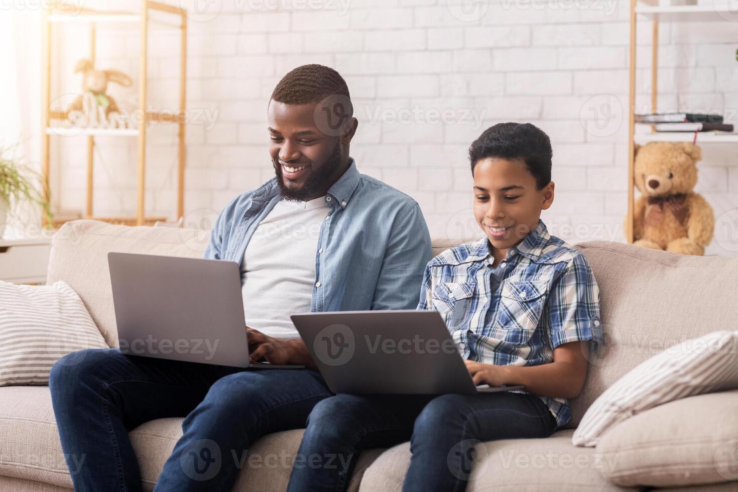 Online Gaming Concept. Happy Afro Father And Son Playing Games On Laptops Together, Sitting With Computer On Laps At Home photo