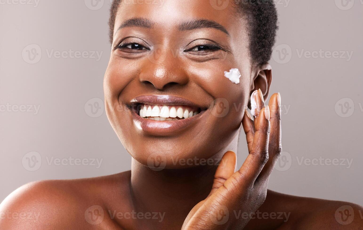 Skin Care Concept. Closeup Portrait Of Happy African American Woman Applying Moisturizing Cream on Face Over Grey Background photo