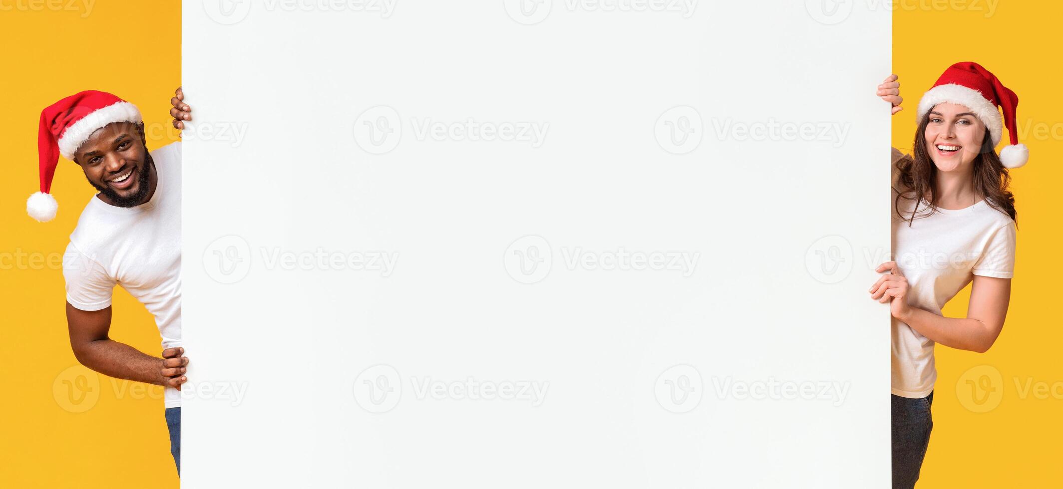 Christmas advertisement. Joyful interracial couple in xmas hats holding white billboard with empty space for your text, yellow background, panorama photo
