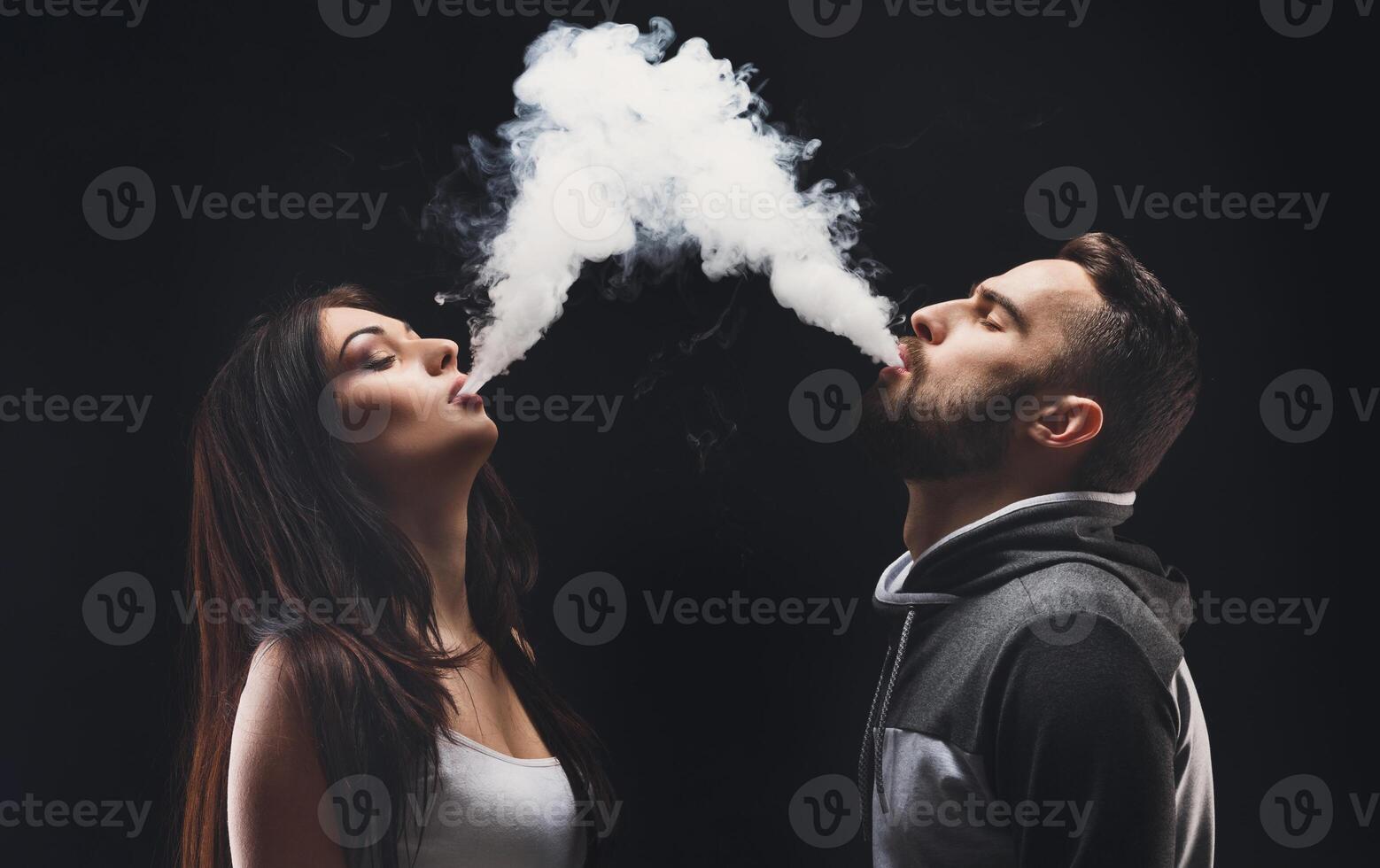 Couple vaping. Young man and woman blowing smoke to join it in one cloud at black studio background. Relationship and vape addiction concept with copy space photo