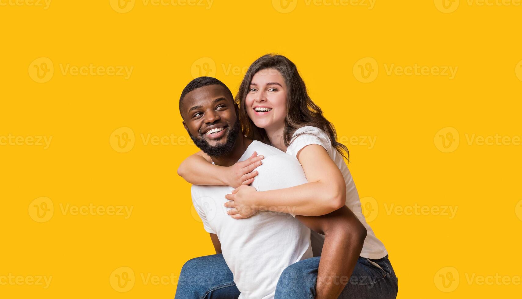 Happy interracial couple. Cheerful black guy piggibacking his joyful girlfriend, having fun together over yellow studio background, empty space photo
