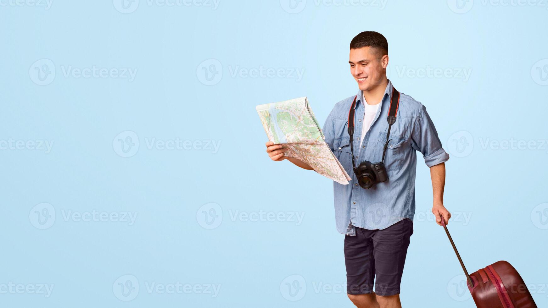 Lost in city. Young male tourist with suitcase holding city map, going towards copy space, orange studio background, panorama photo