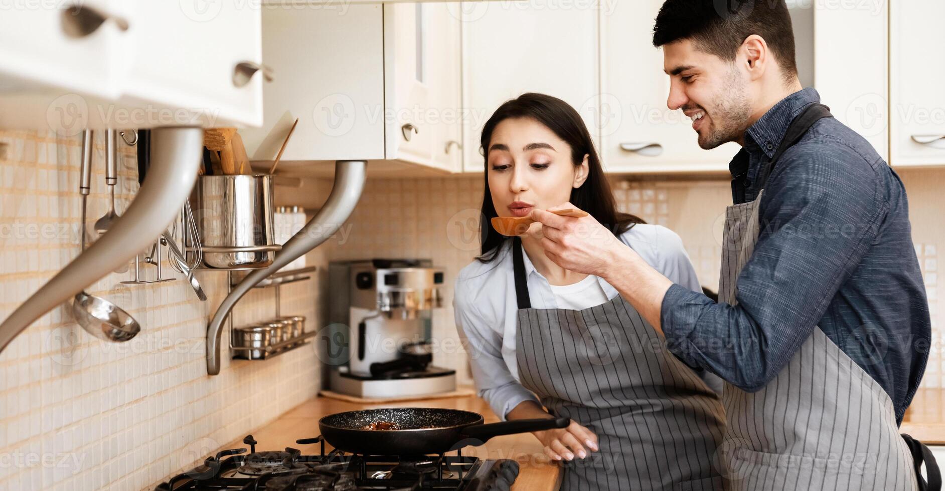 Cooking Together Concept. Smiling man feeding his woman, lady tasting hot fried meal using wooden spoon photo