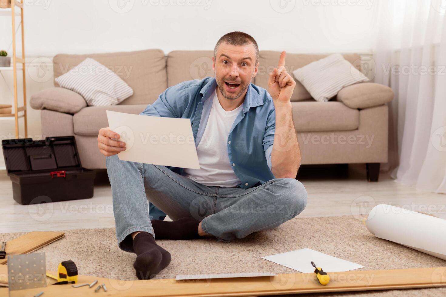 Excited Man Assembling Furniture Pointing Finger Up Having Idea Sitting On Floor At Home, Reading Installation Instruction. Selective Focus photo