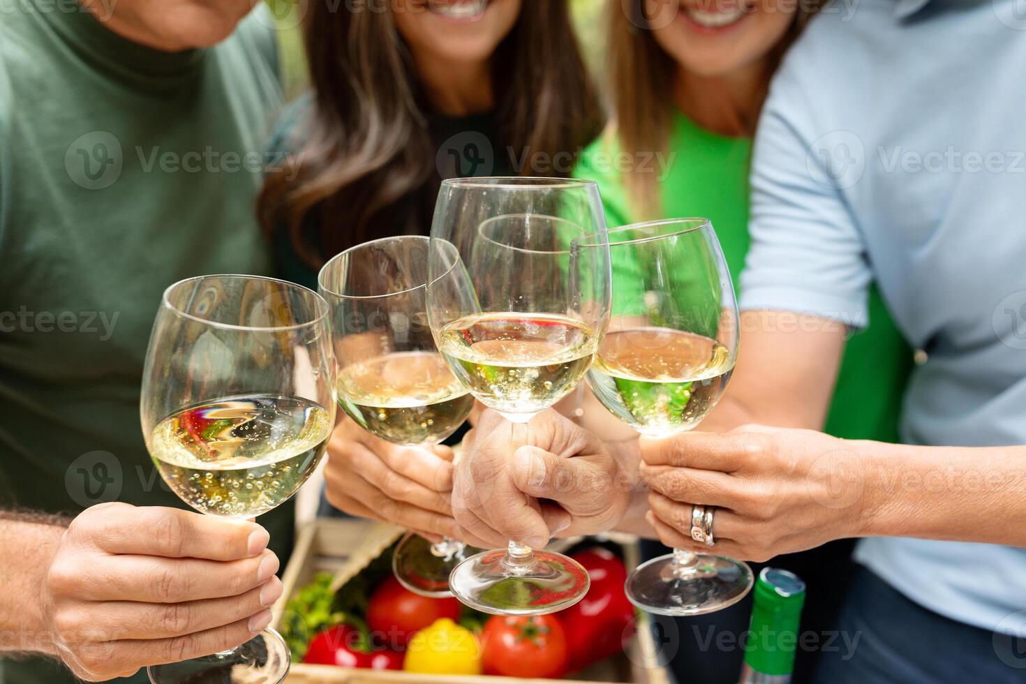A group of friends are gathered outdoors, smiling and raising glasses filled with a light beverage. They enjoy each other's company in a vibrant, green environment surrounded by fresh produce. photo