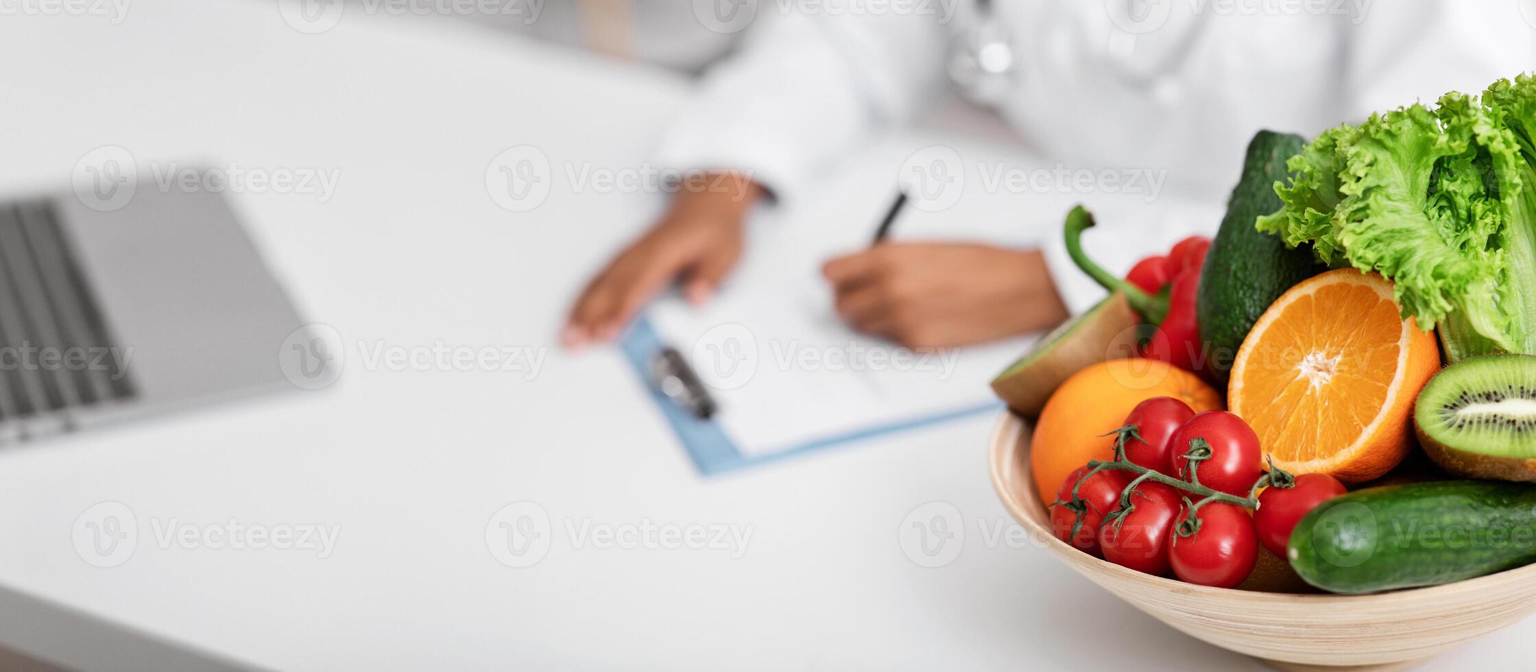 Close up of bowl of fresh fruits and vegetables on dietologist workplace, doctor making prescription photo