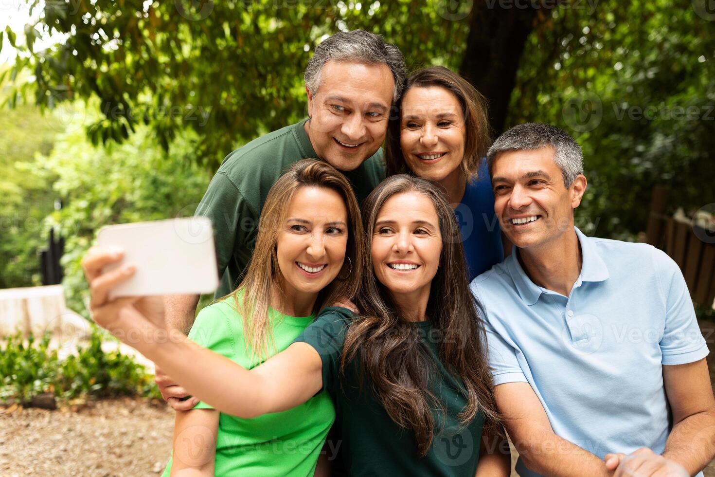 A group of five friends gathers in a park, smiling and posing together for a fun selfie amidst lush greenery on a sunny day. Their joy captures the essence of friendship. photo