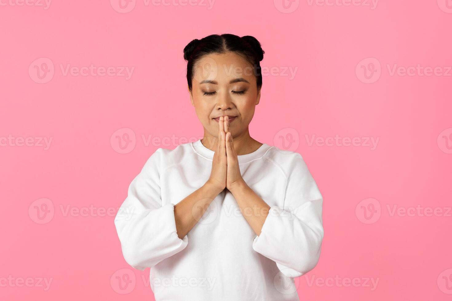 Asian woman is quietly practicing mindfulness with her hands pressed together in a gesture of peace. She stands against a vibrant pink background, displaying tranquility and focus. photo