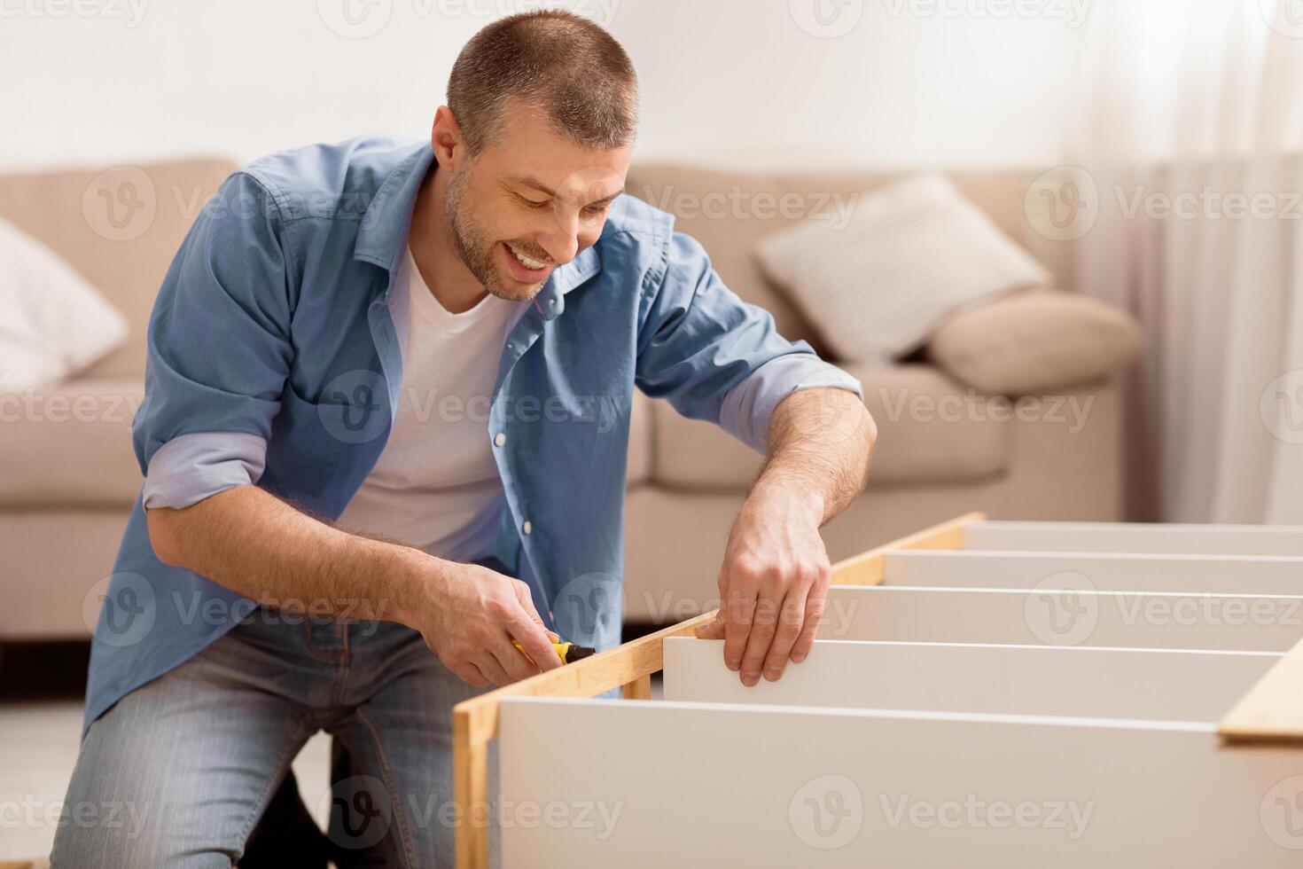 Furniture Repair. Happy Man Fixing Cabinet Using Screwdriver Making Renovation Sitting On Floor At Home. Selective Focus photo