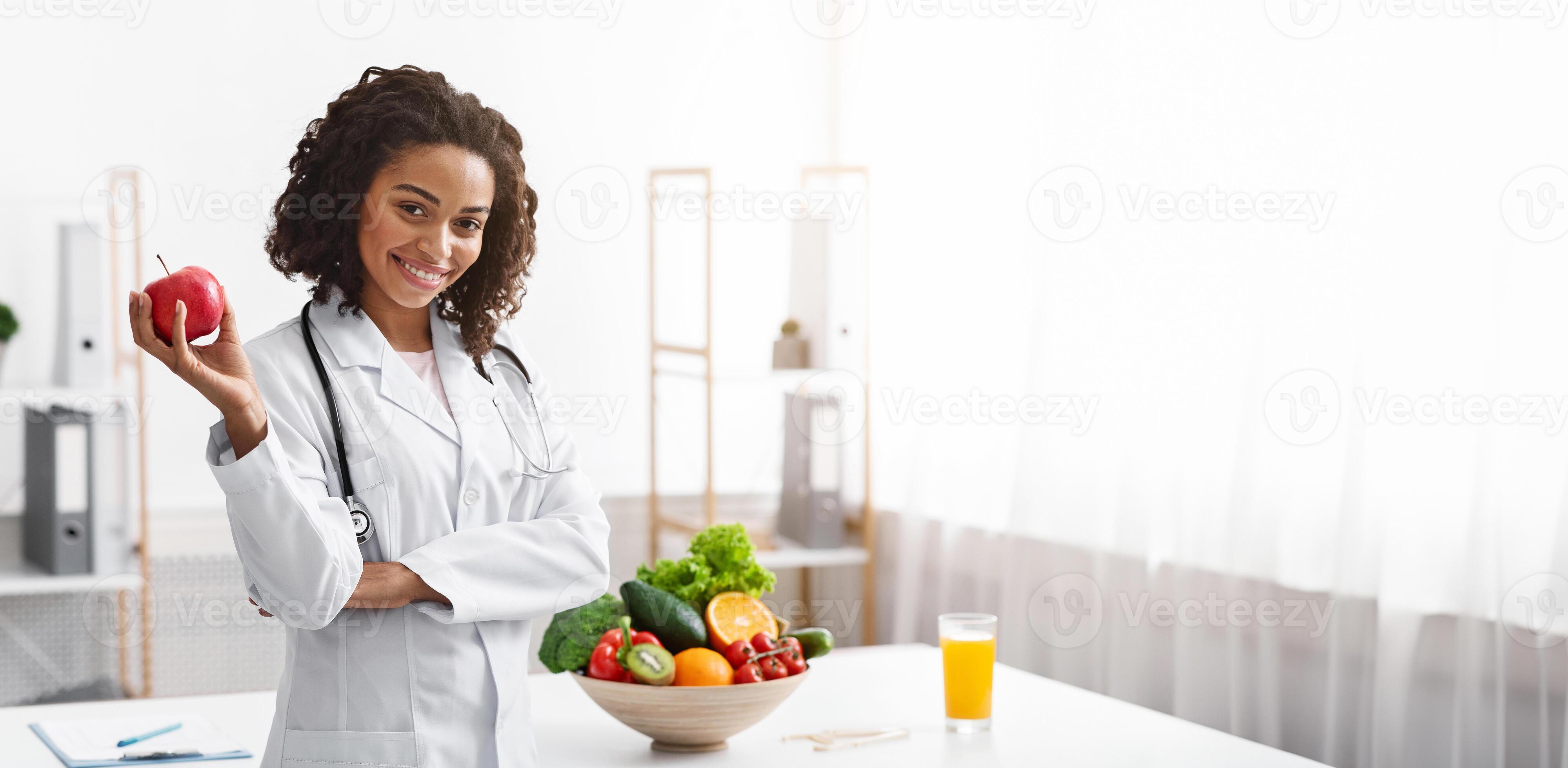 Positive black woman nutritionist posing at clinic holding apple copy Positive black woman nutritionist posing at clinic holding apple copy