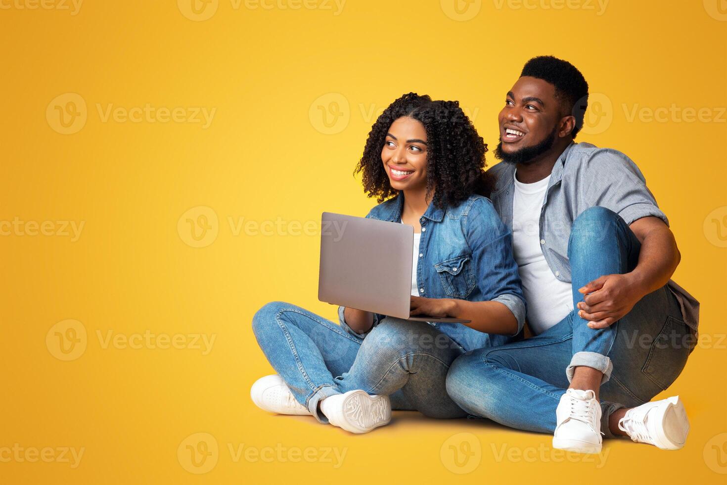 Smiling black couple sitting on floor with laptop and looking aside at copy space on yellow background, panorama photo