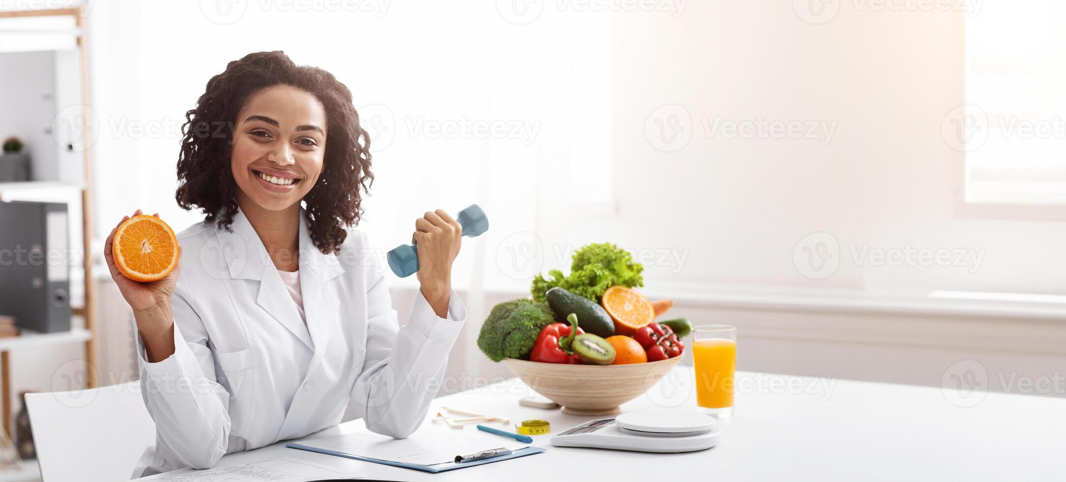 Sport and vitamins for immunity. Happy black woman doctor holding orange and barbell in clinic, posing at her workplace, free space photo