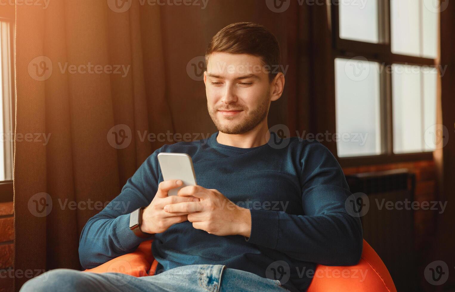 Mobile App. Student Guy Using Phone Texting And Browsing Internet Sitting In Beanbag Chair Indoor. Selective Focus photo