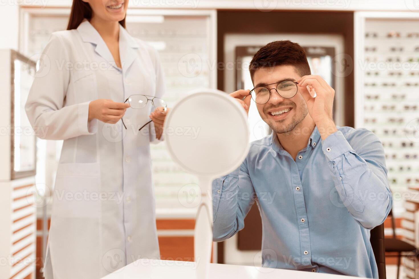 Optical Shop. Lady optician in white coat holding glasses, helping handsome guy to choose the most appropriate eyewear photo