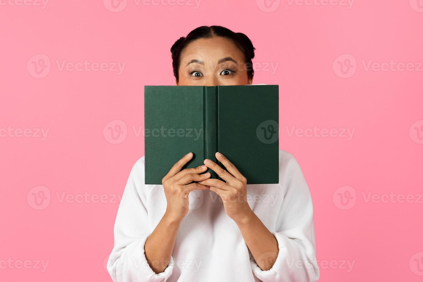 A woman with an excited expression playfully holds a green book in front of her face, against a vibrant pink background, showcasing a moment of joy and surprise. photo