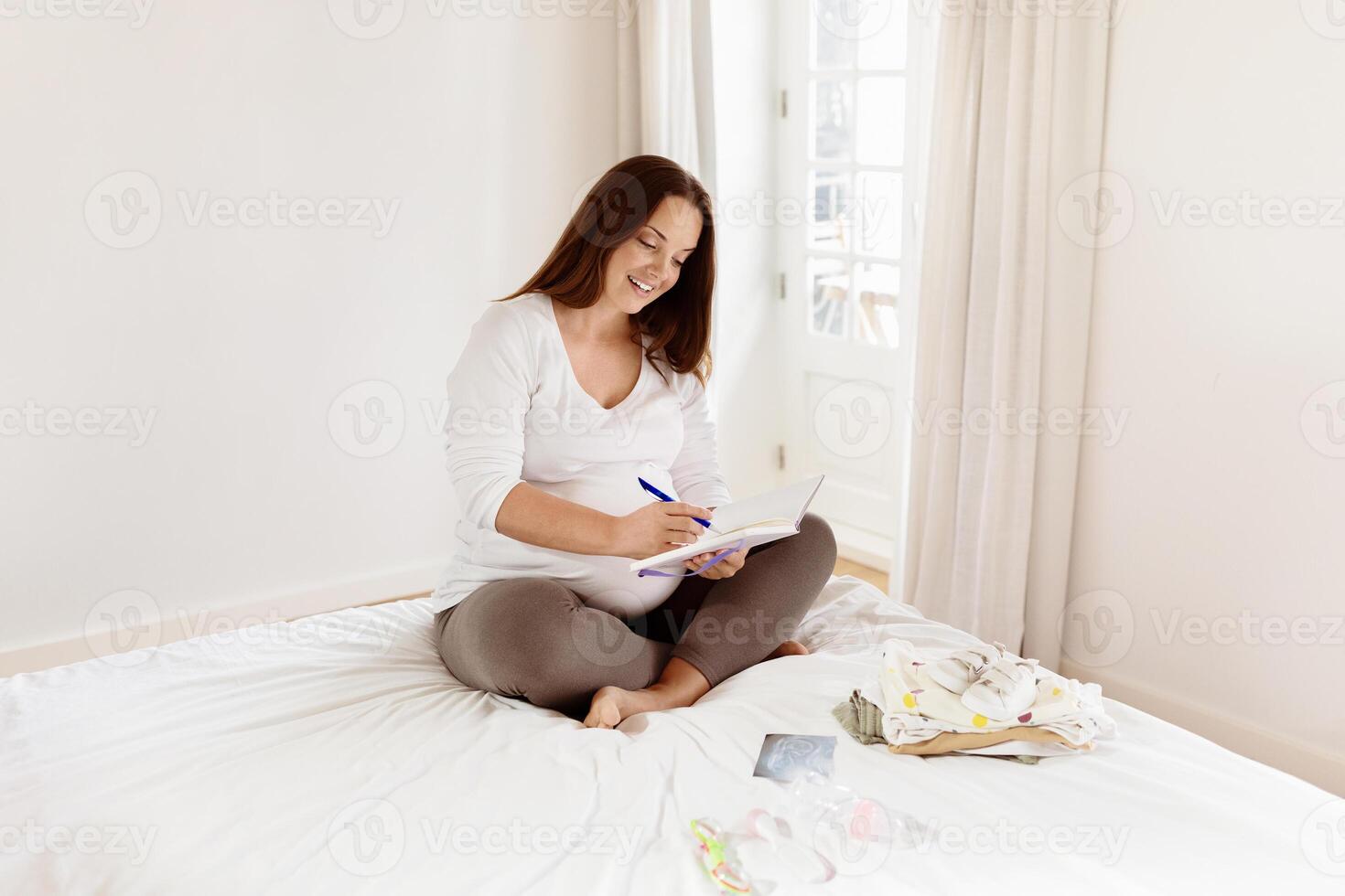 A pregnant woman sits cross-legged on a bed, happily writing in a notebook. She is surrounded by baby clothes and enjoys a moment of creativity in a well-lit, airy room. photo