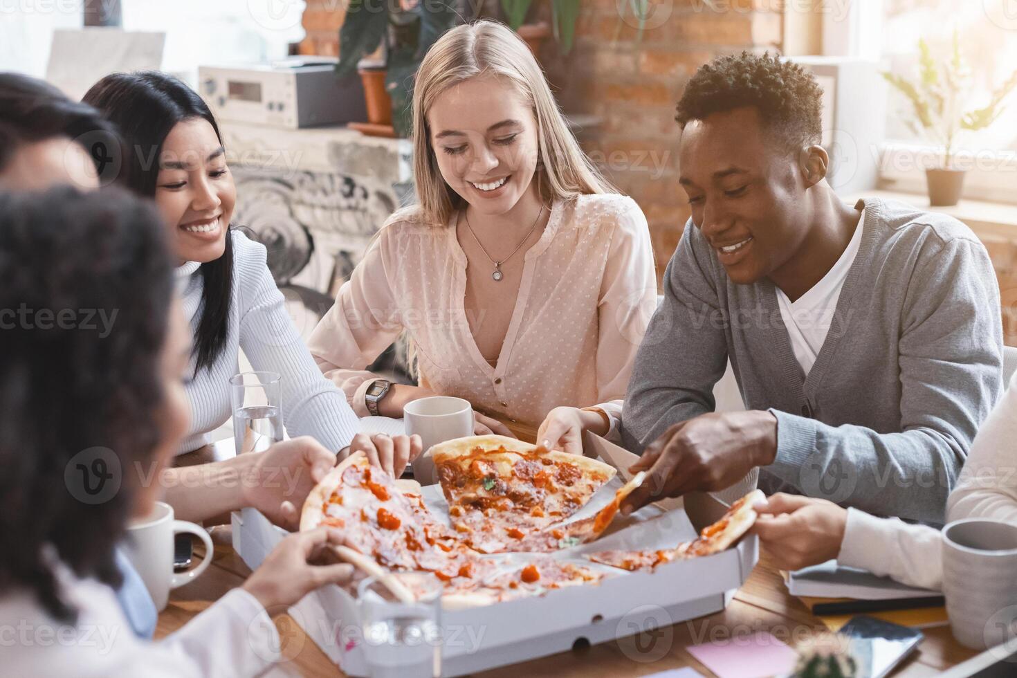 Happy multiracial colleagues eating pizza in office, having break while business meeting photo