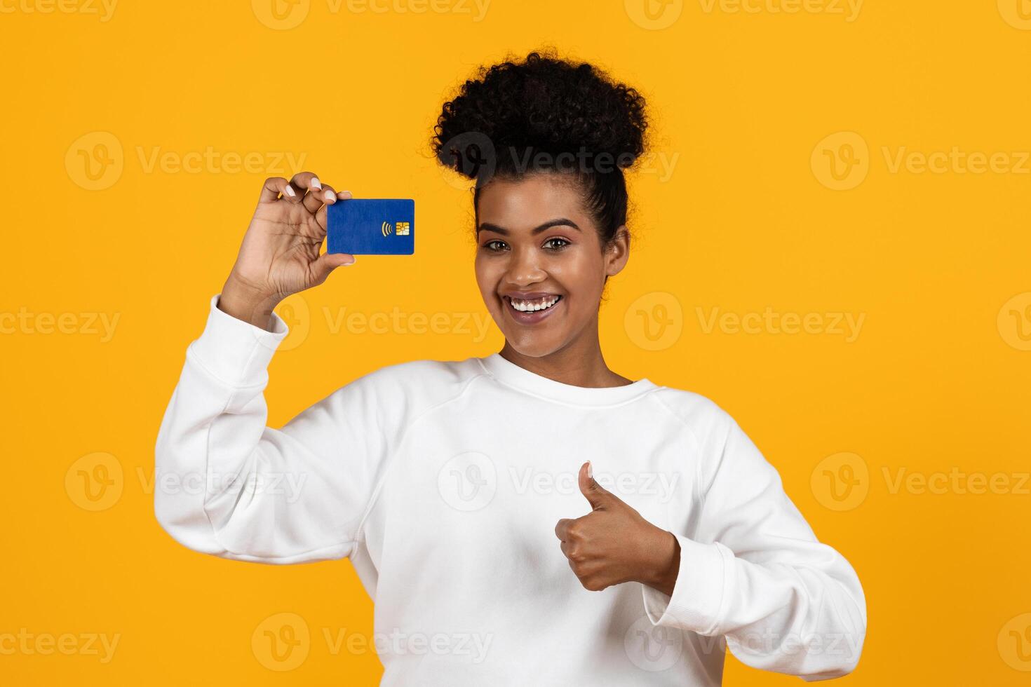 A cheerful young woman displays a card with one hand while giving a thumbs up with the other. She stands against a bright yellow background, radiating positivity and excitement. photo