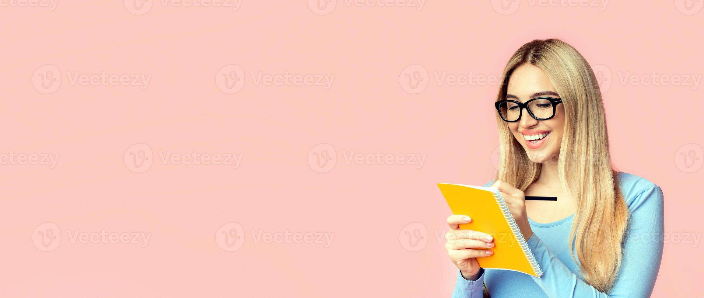 Writing Checklist. Portrait of smiling girl student taking notes in notepad isolated over pink studio wall, copyspace photo