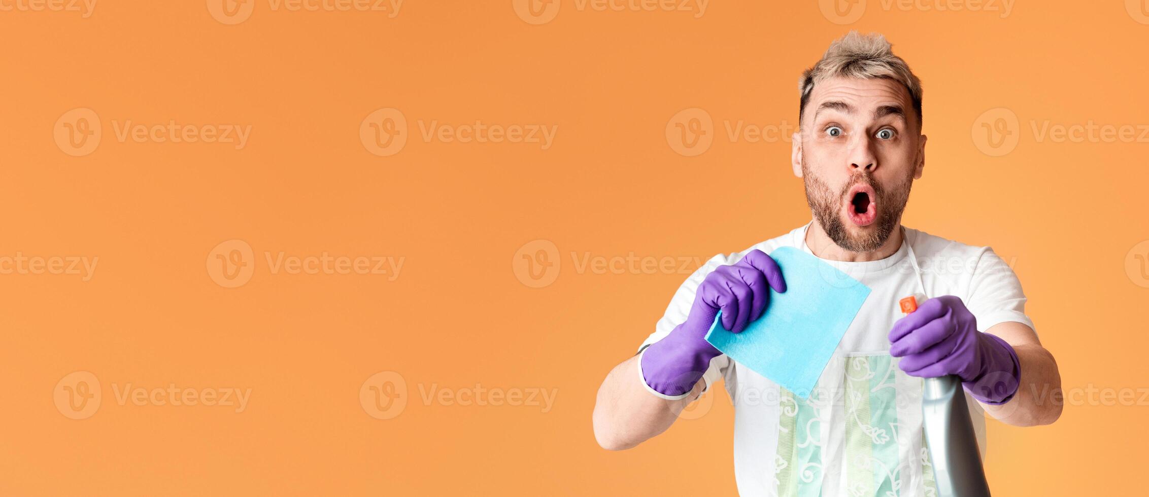 Surprised Young man is cleaning, Holding spray and bast in hands. Isolated on yellow background photo