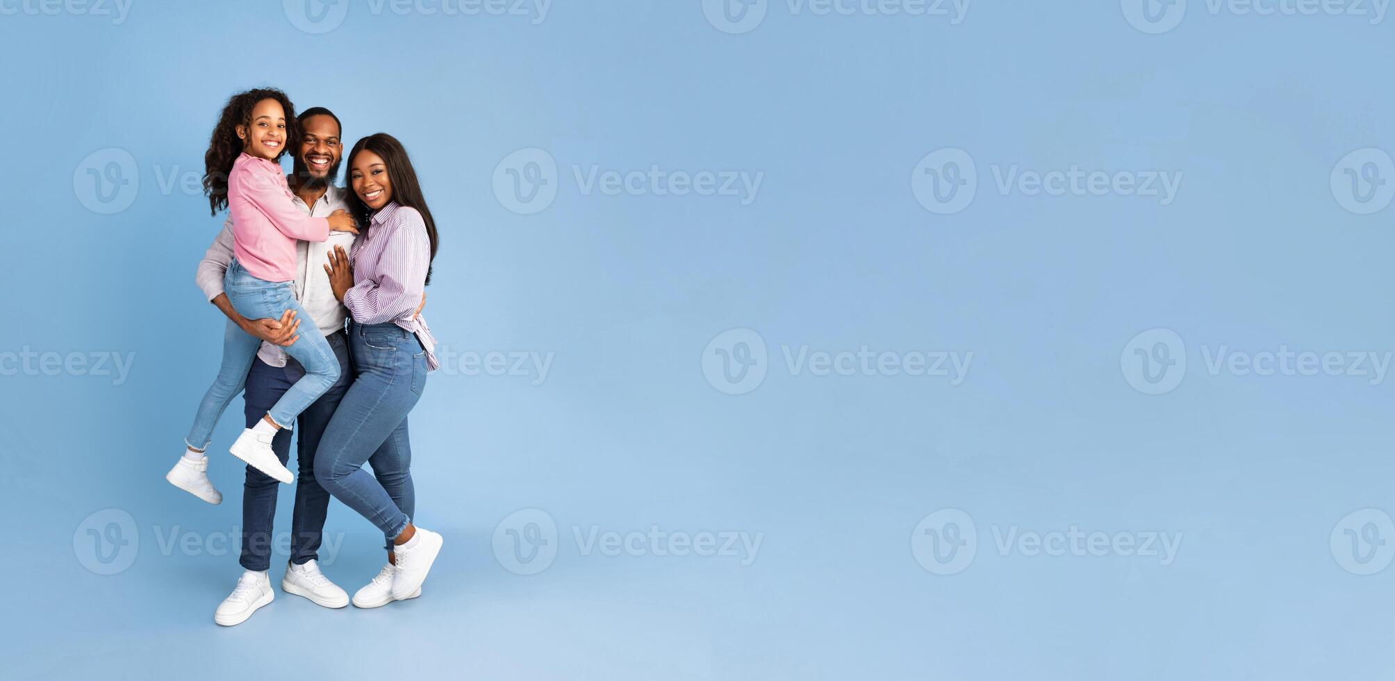 Full Body Length Of Cheerful Black Man Holding Excited Daughter On Hands, Hugging Wife, Looking And Posing At Camera, Standing Isolated Over Blue Studio Background, Panorama, Banner, Free Copy Space photo