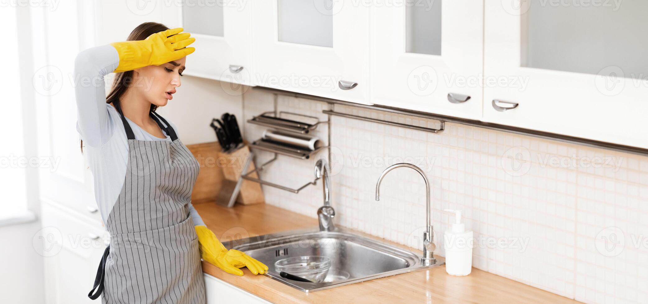 Domestic Life Concept. Tired woman in apron standing in kitchen, looking at dirty dishes in sink, copy space photo