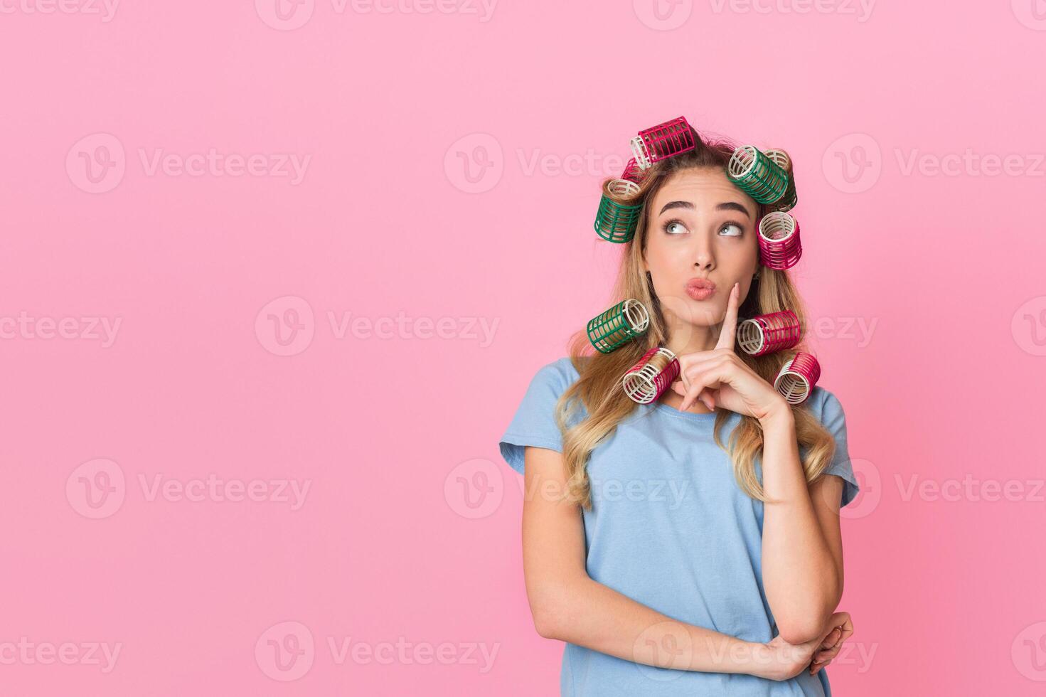 Pensive young housewife in curlers presses finger to cheek and looks up to empty space, isolated on pink background, studio shot photo
