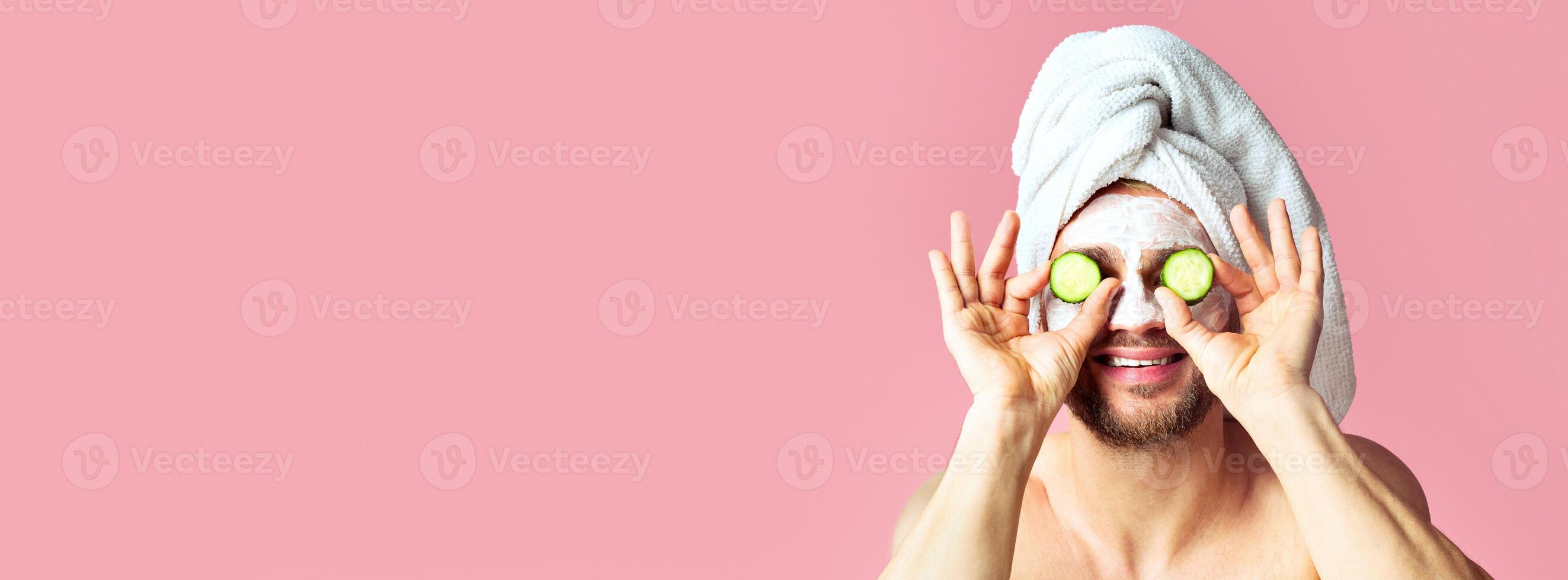 Cheerful guy with mask on his face applies slices of cucumber to eyes, copy space photo