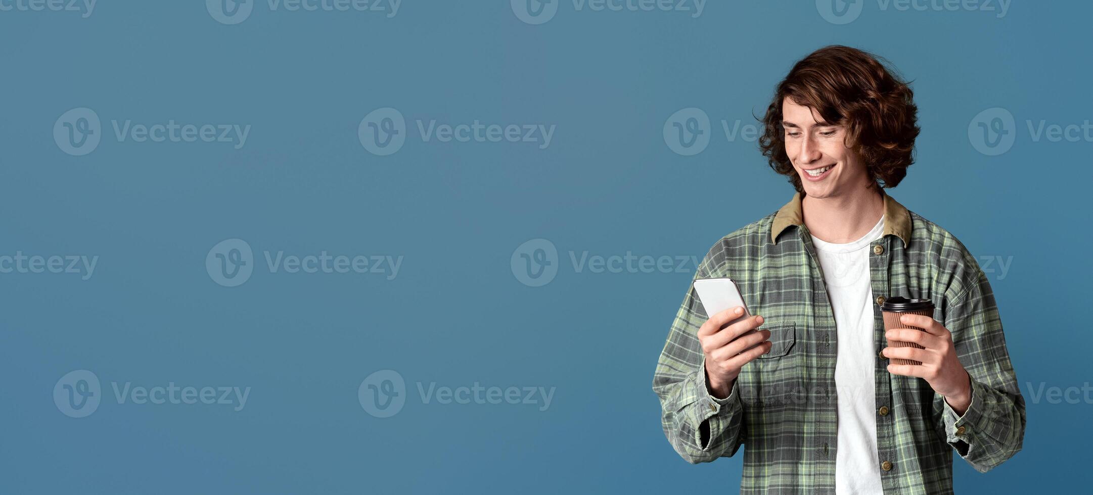 Smiling young guy with smartphone and cup of coffee communicating online via cellphone, turquoise background photo