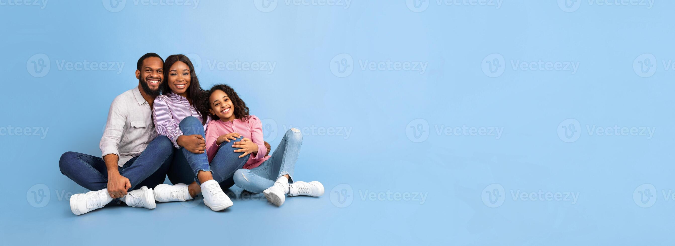 Smiling positive happy milennial African American parents sitting on the floor with teenage adolescent girl, hugging daughter isolated on blue color background, panoramic shot, free copy space, banner photo