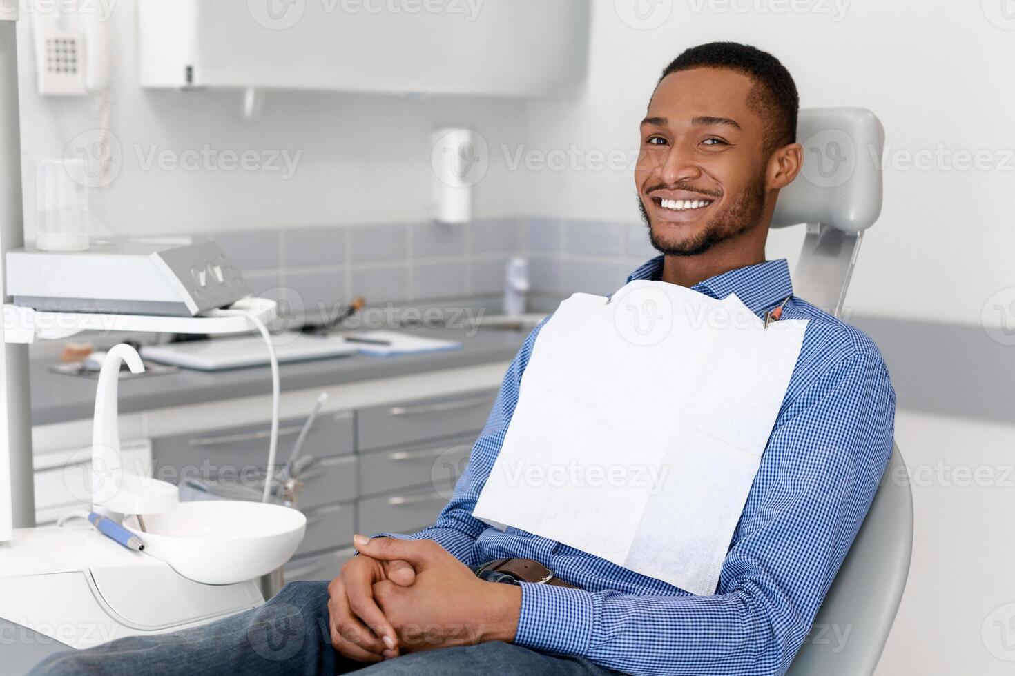 Happy black guy sitting on dentist chair and smiling at camera, attending dental clinic, copy space photo