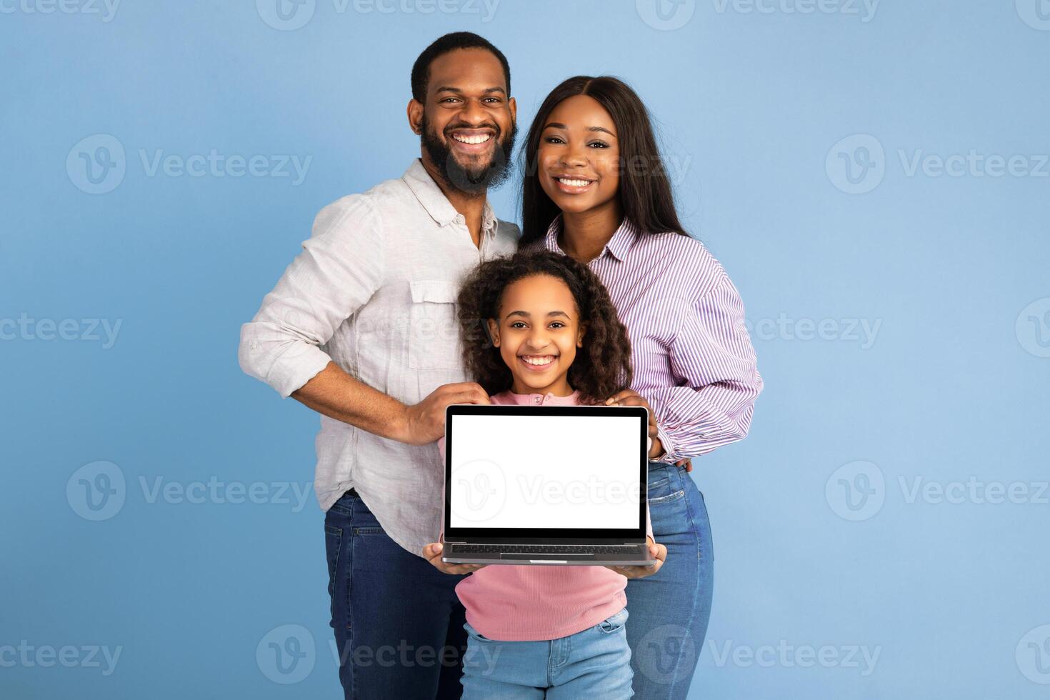 Portrait of happy family of three African American people showing laptop with white empty screen, smiling girl holding gadget with blank space for mock up, standing over blue studio background photo