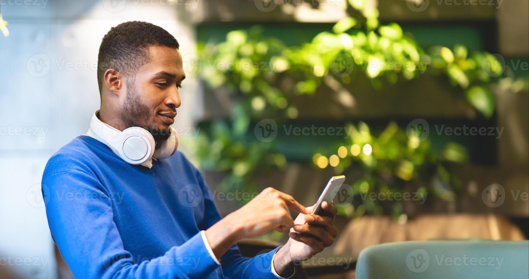 Technology Concept. Side view of young afro student in headphones using smartphone sitting at modern cafe photo