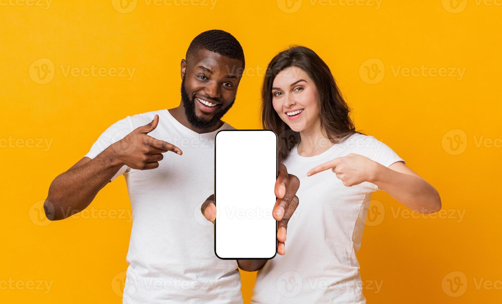 Great app.Happy multiracial couple holding and pointing at smartphone with black screen for mockup, standing together over yellow background photo